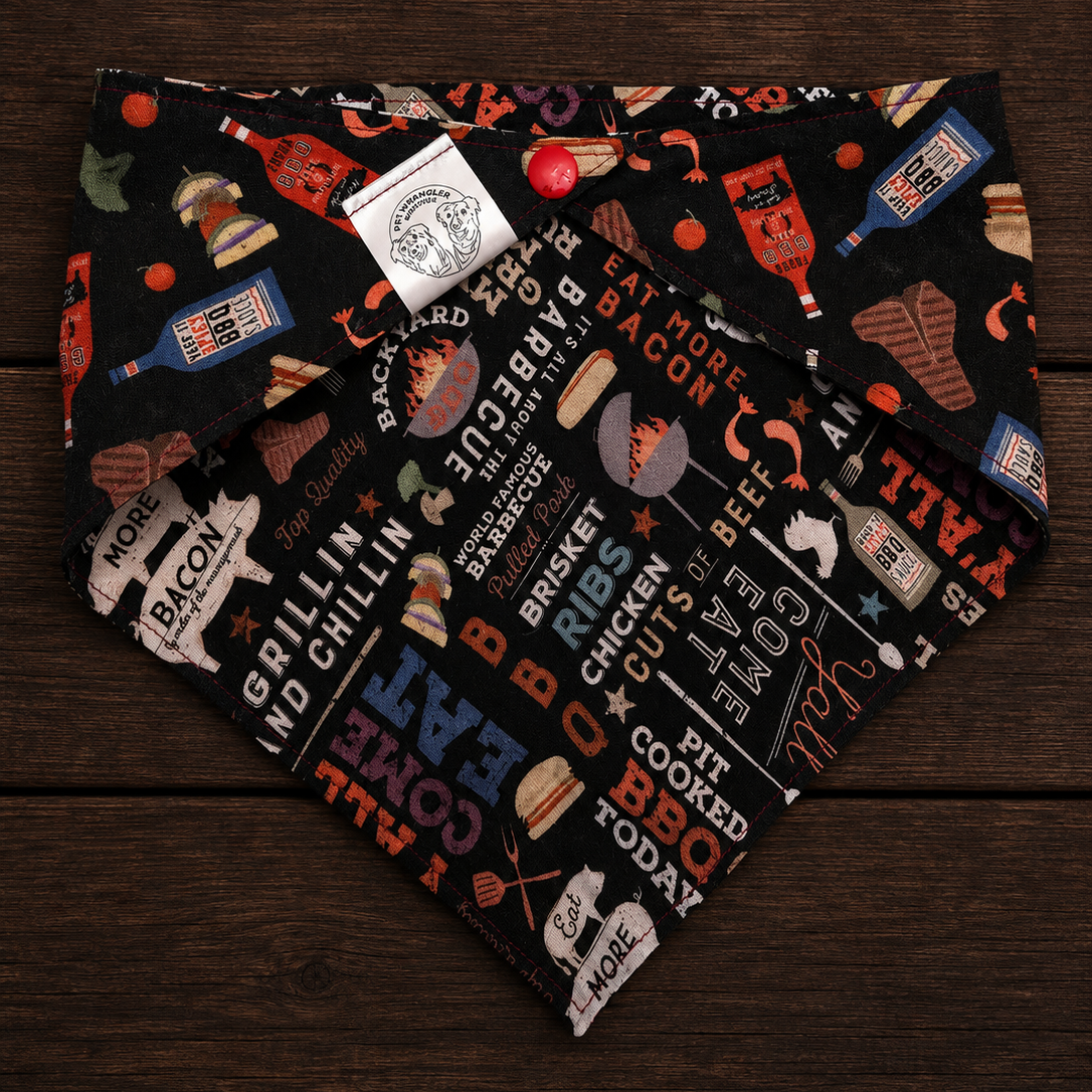 A black bandana dog collar with a colorful barbecue-themed graphic print, laid flat on a dark wood background. The print features illustrations of burgers, hot dogs, bottles, and text like 'BBQ RECEPE', 'BACON', and 'COMFORT FUEL'.